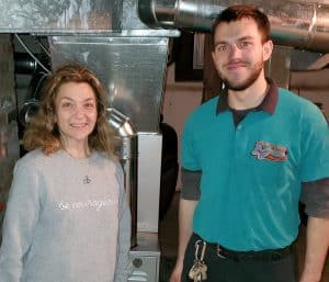 A woman and a man stand indoors next to HVAC equipment. The man wears a teal work shirt and key ring; the woman is in a gray sweatshirt. Both are facing the camera and smiling.