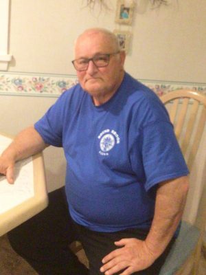 An older man with glasses wearing a blue t-shirt sits at a table in a kitchen, looking at the camera.