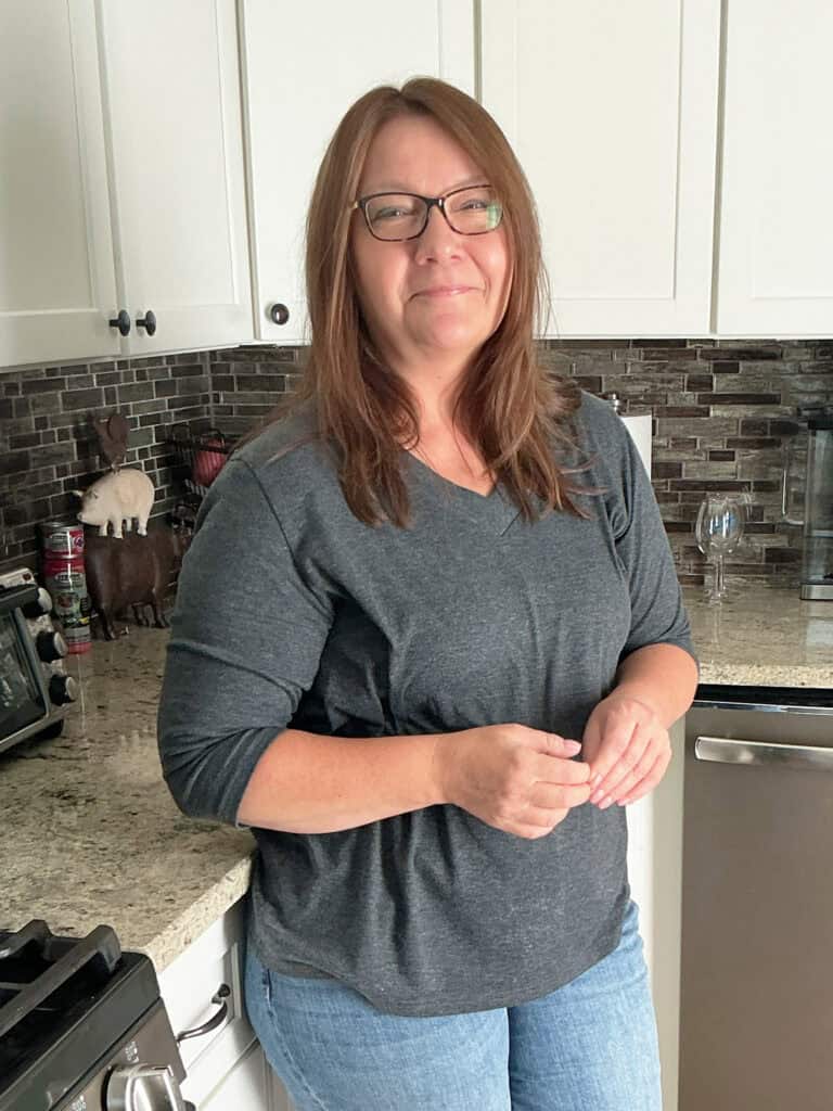 A woman with glasses stands in a kitchen, wearing a dark grey top and blue jeans, with a slight smile on her face.