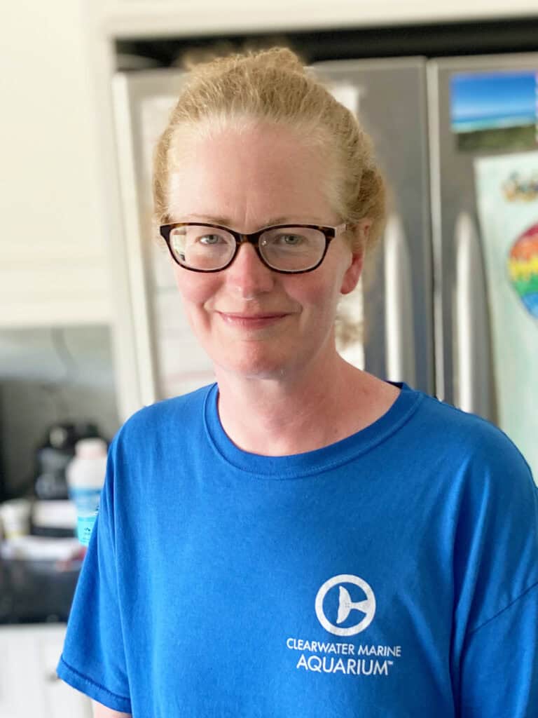 Person with glasses and light hair pulled back, wearing a blue Clearwater Marine Aquarium t-shirt, stands indoors in front of a refrigerator.