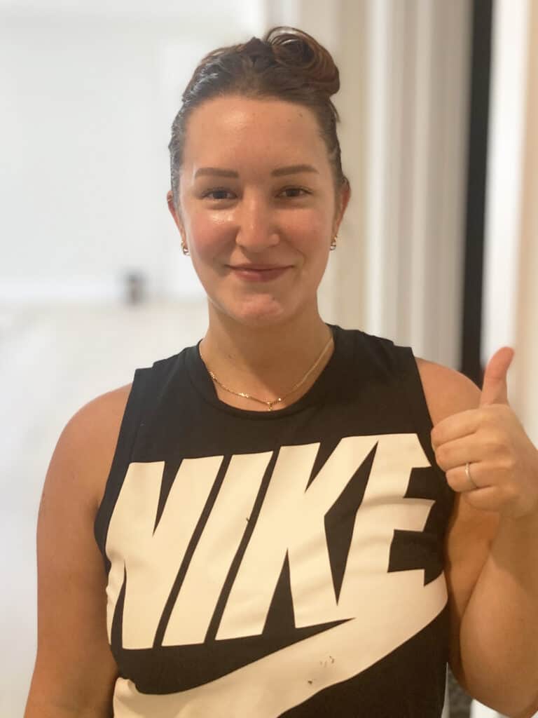 A woman wearing a black Nike tank top gives a thumbs up and smiles at the camera indoors.