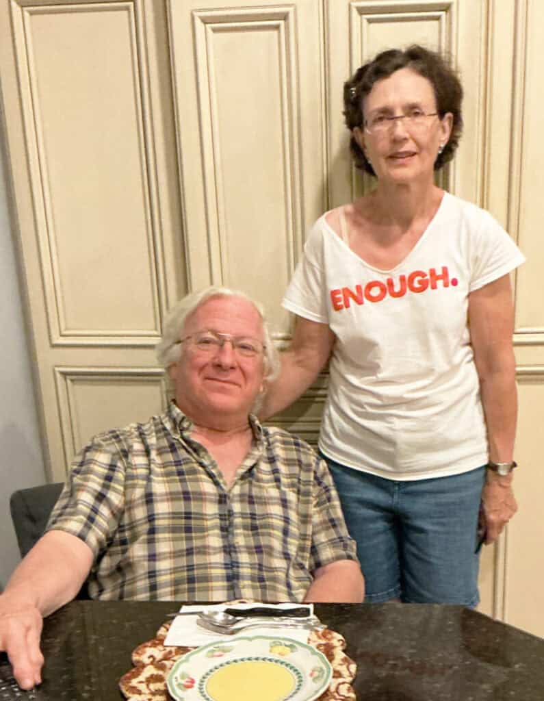An older man sits at a table with a plate and bowl, while an older woman stands beside him wearing a white shirt that says "ENOUGH." They are indoors in a kitchen.