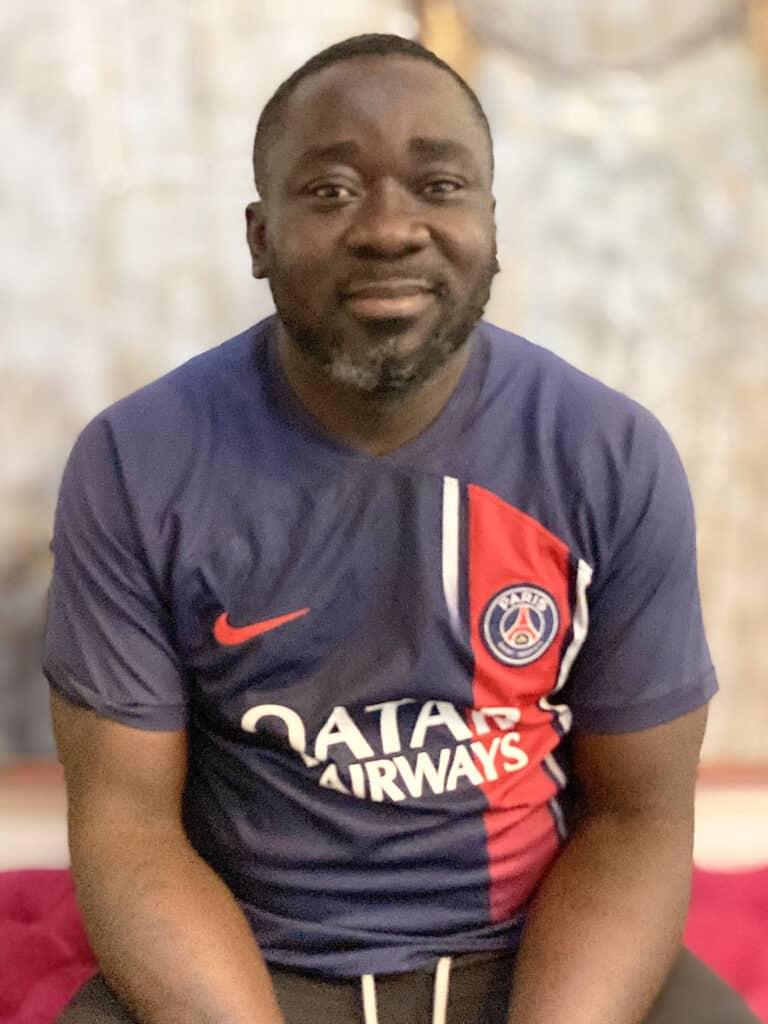 A man with a short beard and mustache is sitting and wearing a navy blue Paris Saint-Germain football jersey.