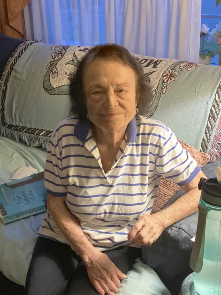An older woman wearing a striped polo shirt sits on a couch with a book beside her and a water bottle in the foreground.