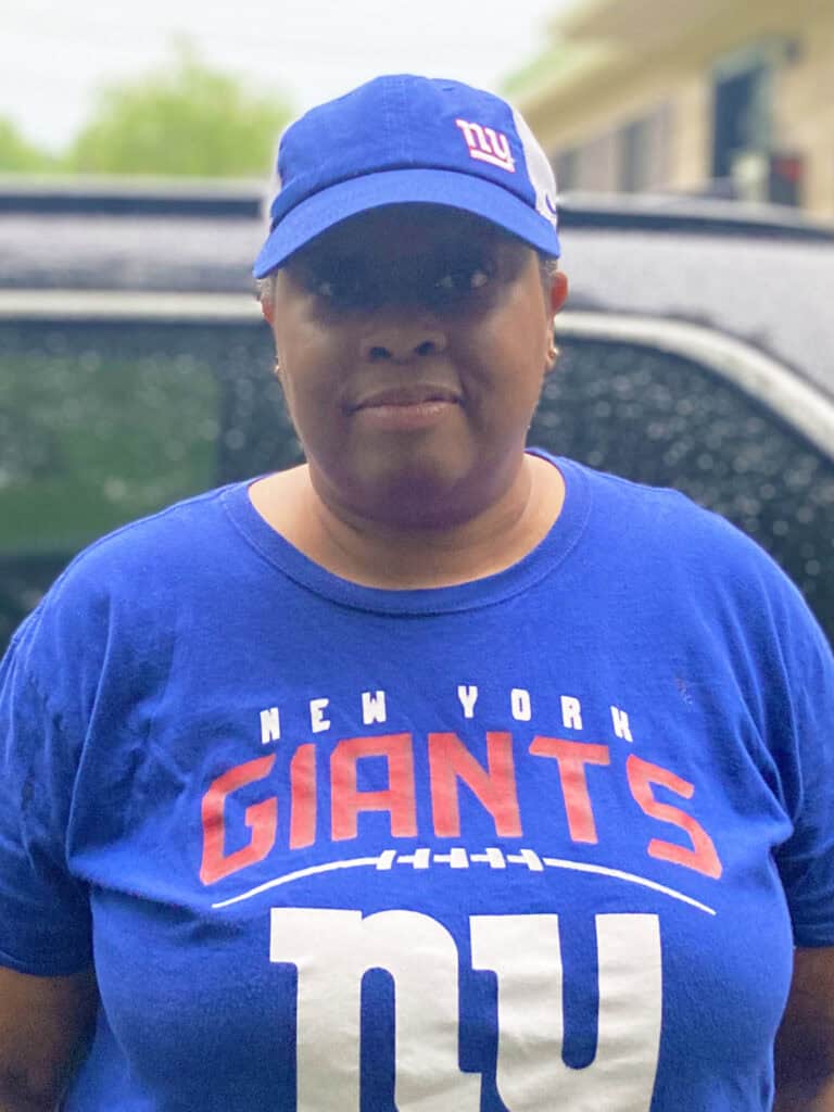 A person wearing a blue New York Giants shirt and matching cap stands outside in front of a car on a rainy day.