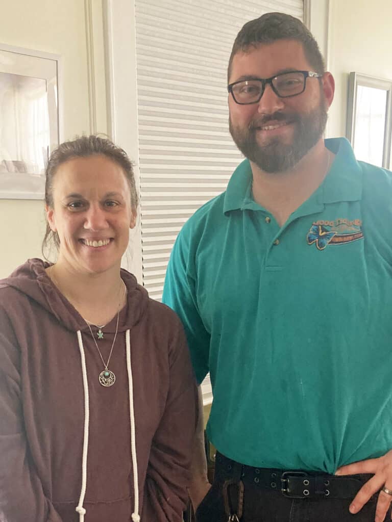 A woman in a maroon hoodie stands next to a man in a teal polo shirt and glasses, both smiling at the camera indoors.