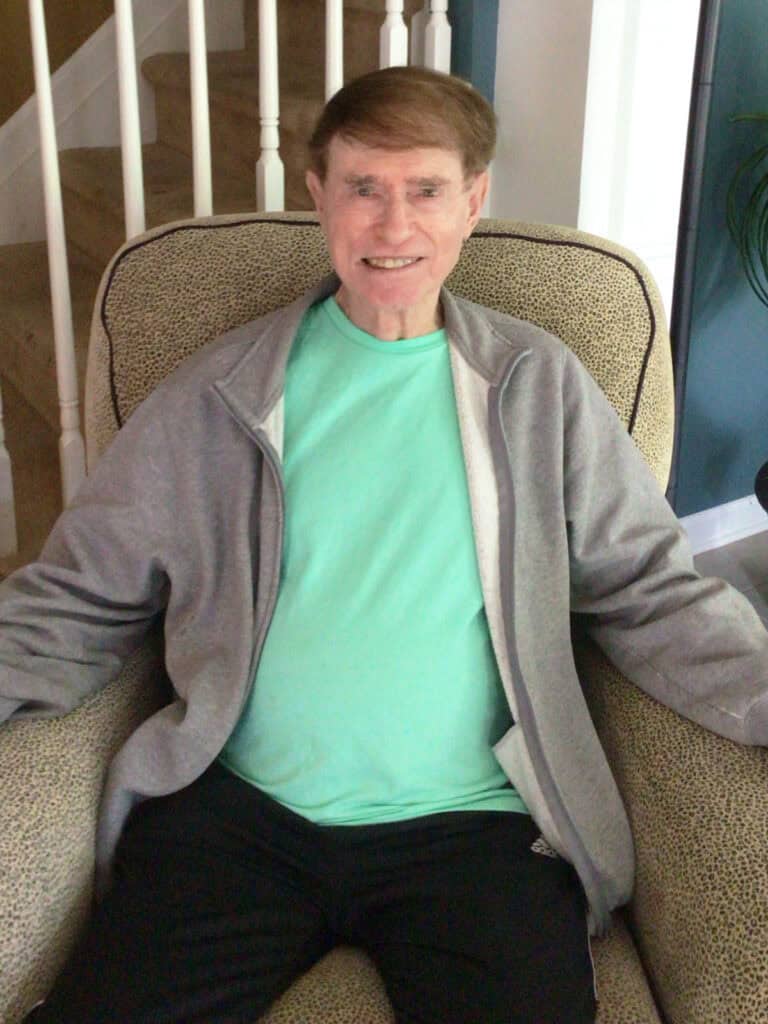 An older man with short brown hair sits in an armchair, wearing a light green shirt, gray zip-up jacket, and black pants, smiling at the camera.