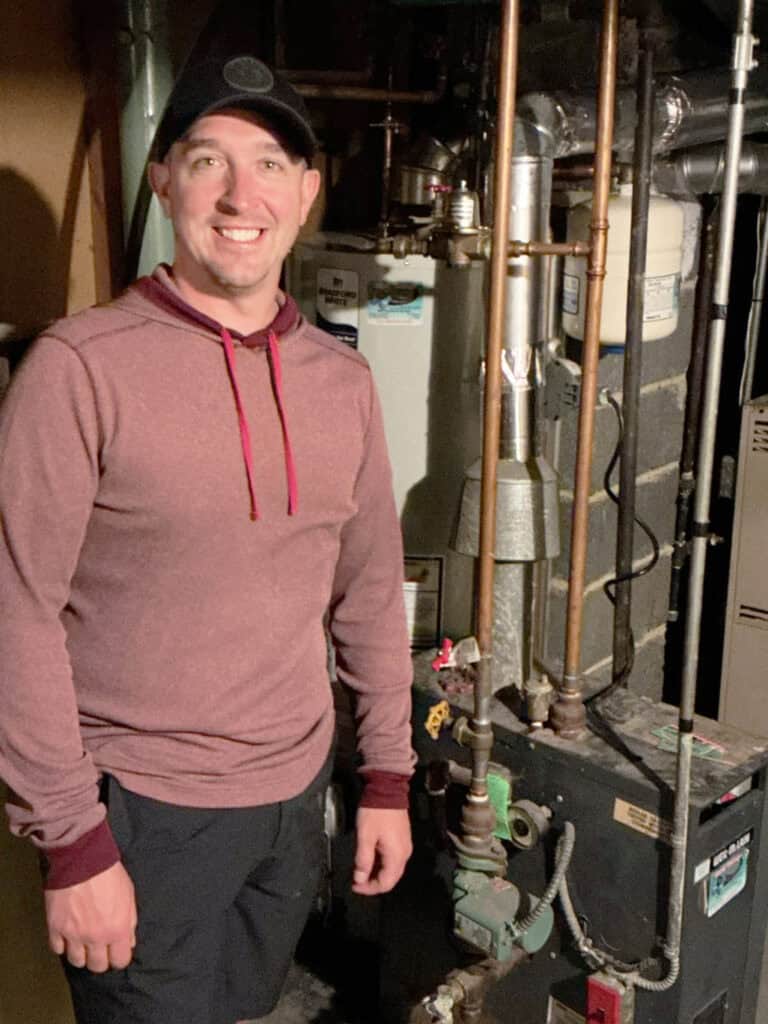 A person wearing a maroon hoodie and black cap stands next to a furnace system in a basement, with pipes and equipment visible in the background.