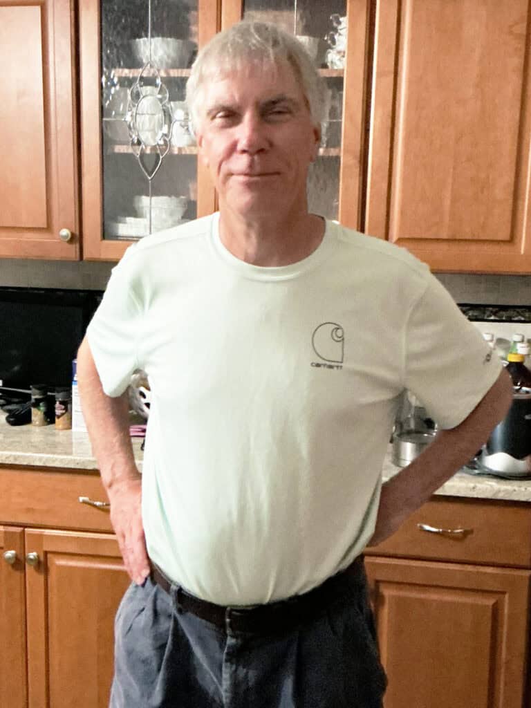A man with gray hair stands in a kitchen with wooden cabinets, wearing a light-colored t-shirt and dark pants, with his hands on his hips.