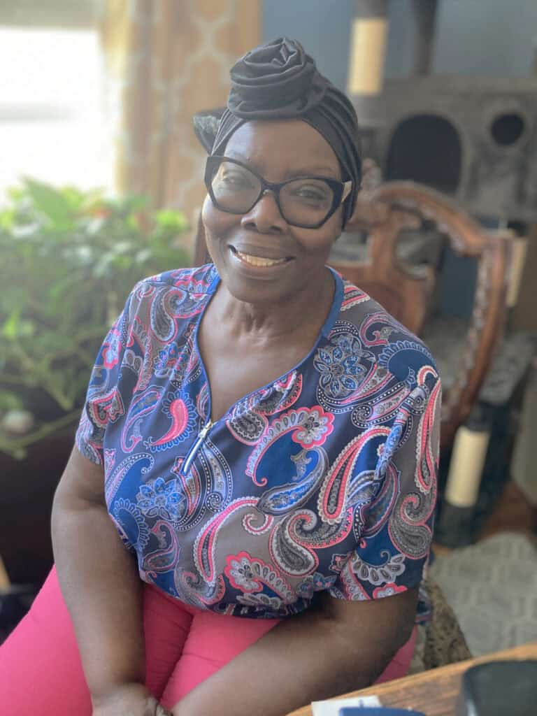 A woman wearing glasses, a patterned shirt, and a headwrap sits indoors, smiling at the camera. There is a plant and furniture in the background.