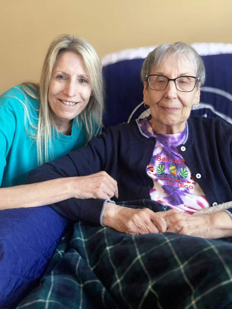 Two women sit together on a bed; one is elderly and wearing glasses, while the other, younger woman smiles and leans on her arm.