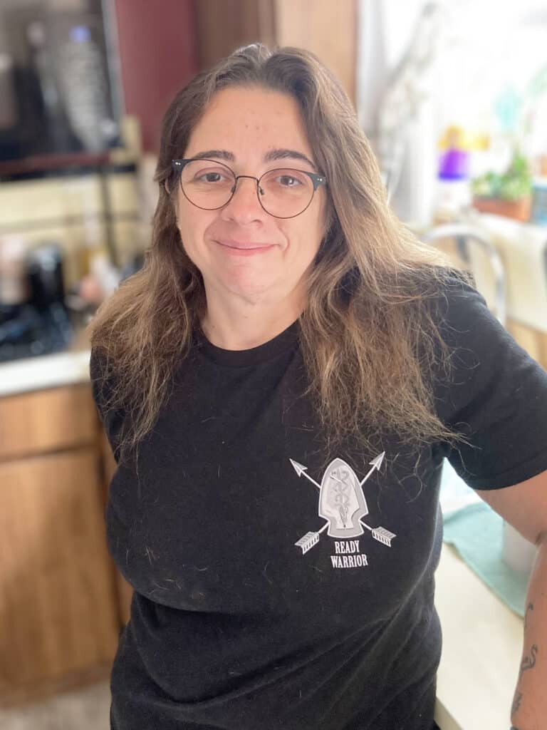 A person with long brown hair and glasses stands in a kitchen, wearing a black "Ready Warrior" t-shirt and smiling at the camera.