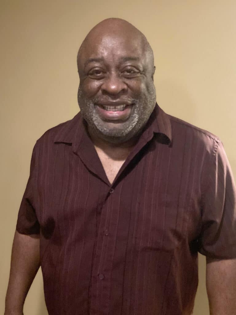 A man with a beard and mustache, wearing a short-sleeved maroon shirt, stands and smiles in front of a plain beige wall.