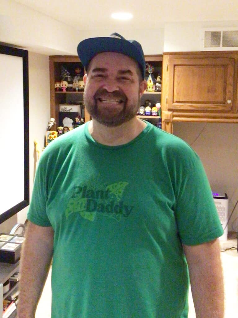 A man wearing a green "Plant Daddy" t-shirt and a blue cap stands smiling in a room with shelves and electronics in the background.