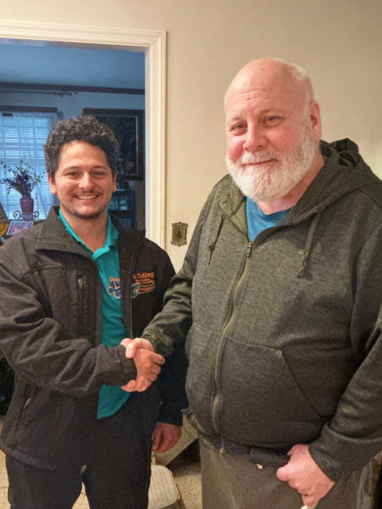 Two men standing indoors shake hands and smile at the camera, one wearing a jacket and the other in a zip-up hoodie.