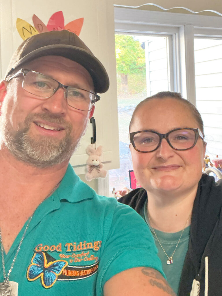Two people wearing glasses smile at the camera indoors. The man wears a teal shirt with a company logo; the woman wears a dark jacket and necklace. A window and decorations are in the background.