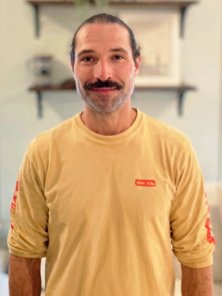 A man with dark hair and a mustache stands indoors wearing a light yellow long-sleeve shirt, with blurred shelves and decor in the background.