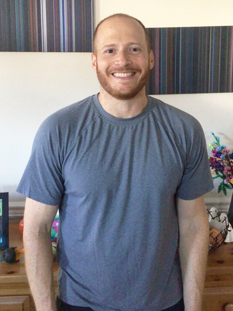 A smiling man with a beard and short hair stands indoors, wearing a gray t-shirt. Colorful decorations and framed pictures are visible on the wooden surface behind him.