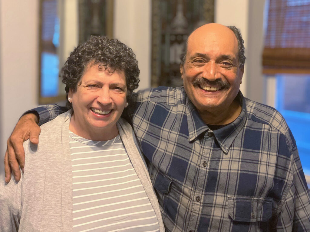 Two older adults, a woman in a light sweater and a man in a plaid shirt, stand indoors smiling with the man's arm around the woman's shoulders.