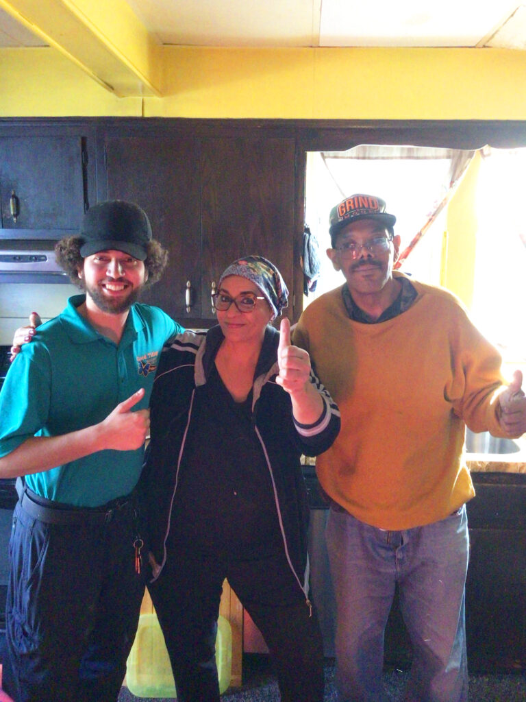 Three adults stand indoors posing for the camera; the person on the left points, the person in the middle gives a thumbs-up, and the person on the right gives a thumbs-up.