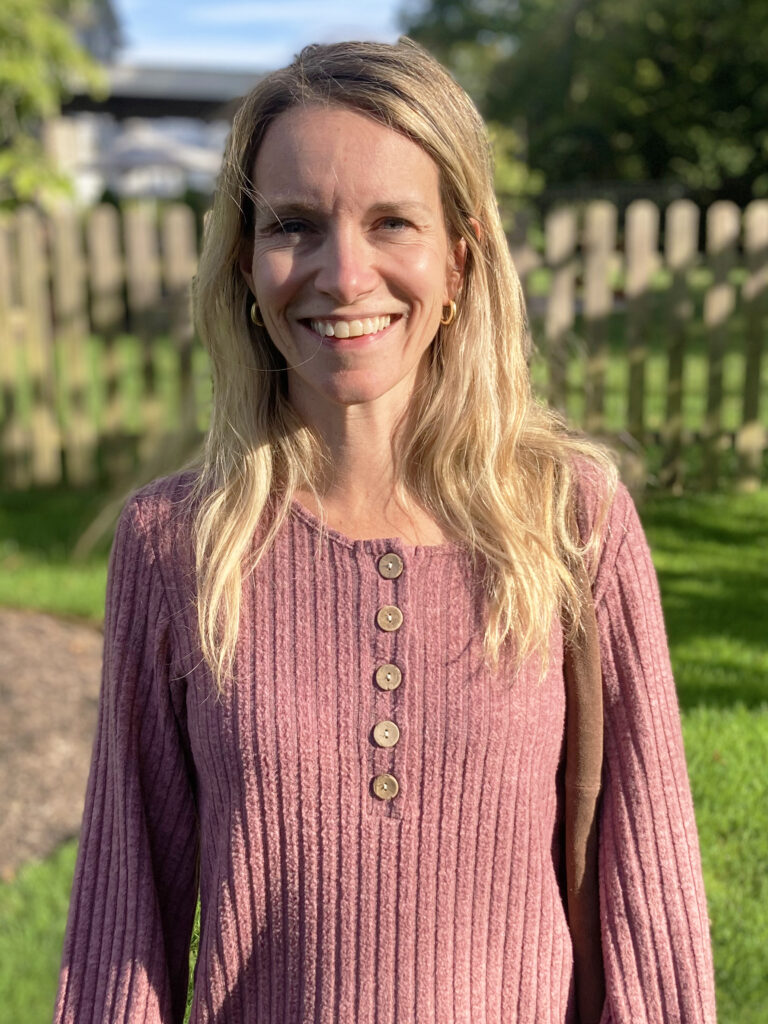 A woman with long blonde hair wearing a ribbed, mauve long-sleeve top stands outside in sunlight near a wooden fence and green grass.