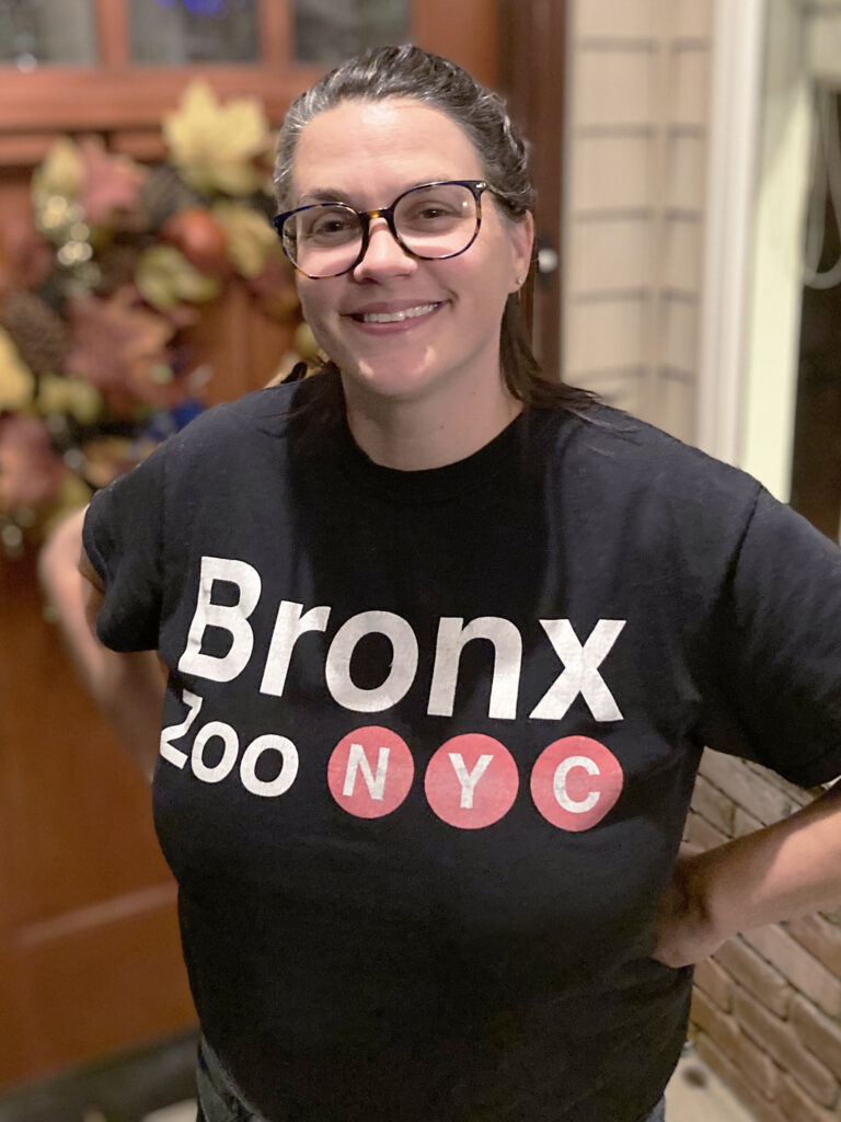 A person with glasses and a ponytail stands indoors, smiling, wearing a black "Bronx Zoo NYC" t-shirt. A decorated door with autumn-themed wreath is in the background.