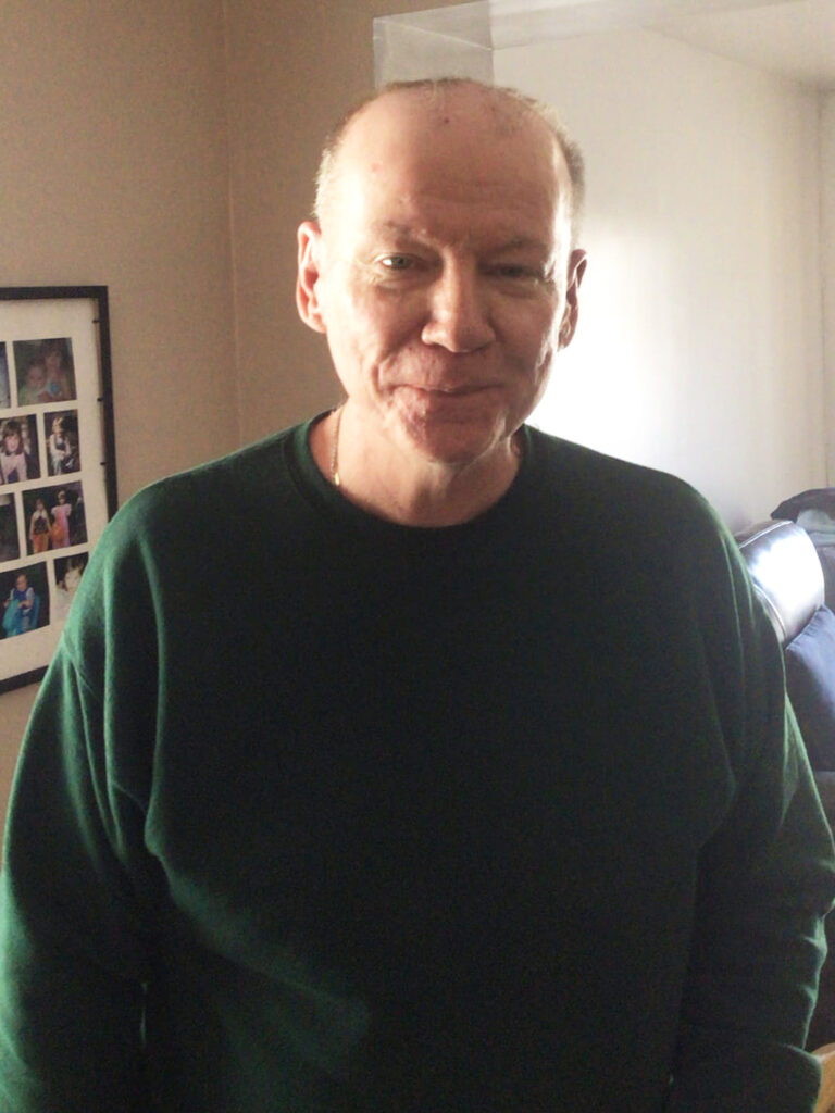 A middle-aged man with short hair wearing a dark green sweater stands indoors, smiling gently at the camera. Framed photos are visible on the wall behind him.