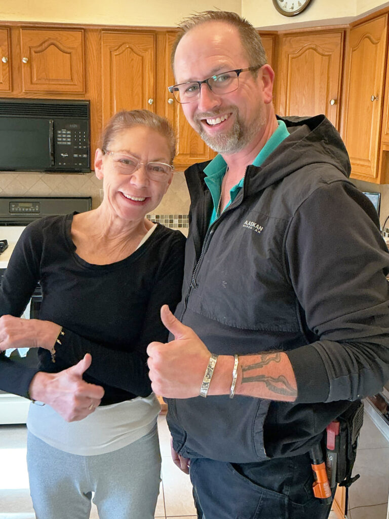 A woman and a man stand side by side in a kitchen, both smiling and giving a thumbs-up gesture. The man wears a toolbelt and vest; wooden cabinets and appliances are visible behind them.
