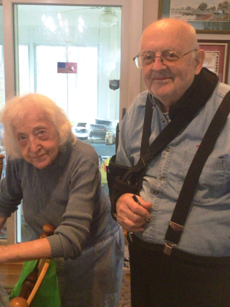 An elderly woman and man stand indoors. The woman leans on a chair, and the man wears glasses, a sling, and suspenders. Both are dressed in casual clothes.