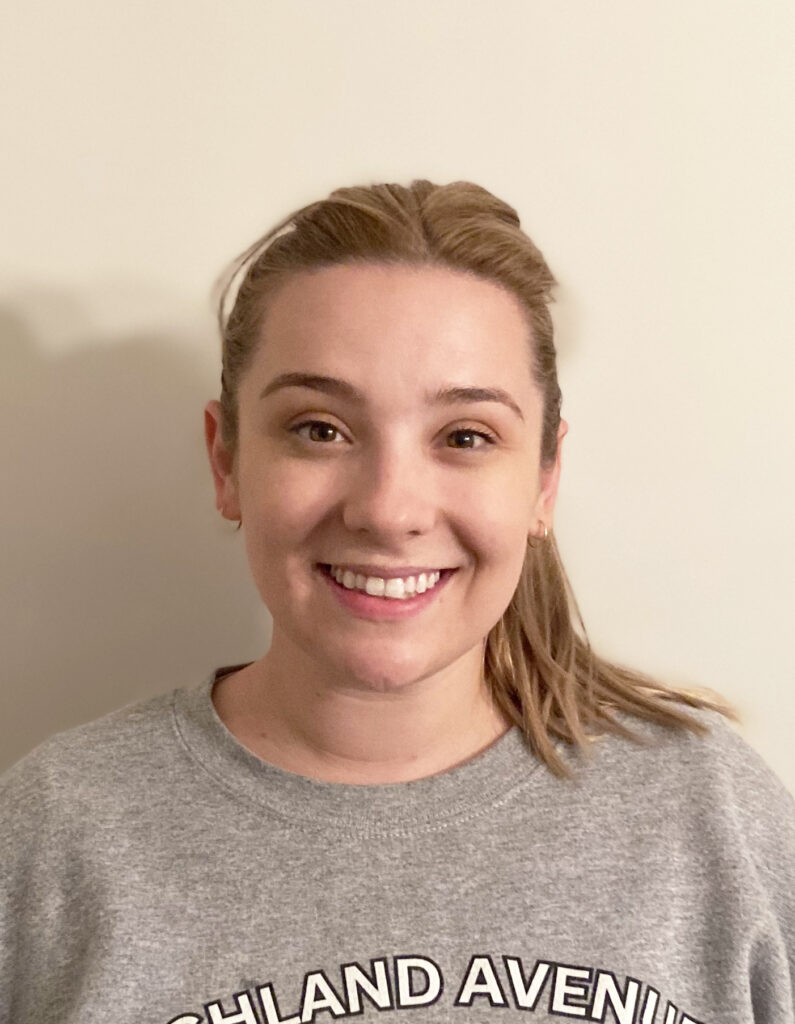 A woman with light brown hair in a ponytail, wearing a grey sweatshirt, smiles in front of a plain off-white wall.