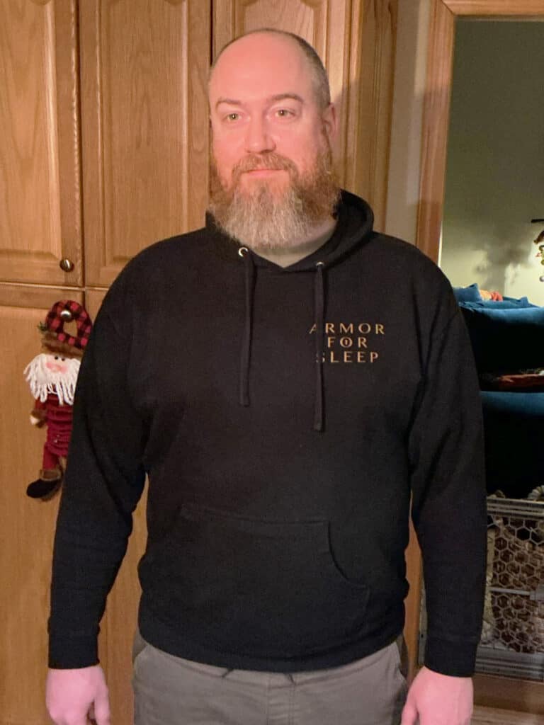 A man with a beard stands indoors wearing a black hoodie that says "ARMOR FOR SLEEP." There is wooden cabinetry and a small plush decoration in the background.