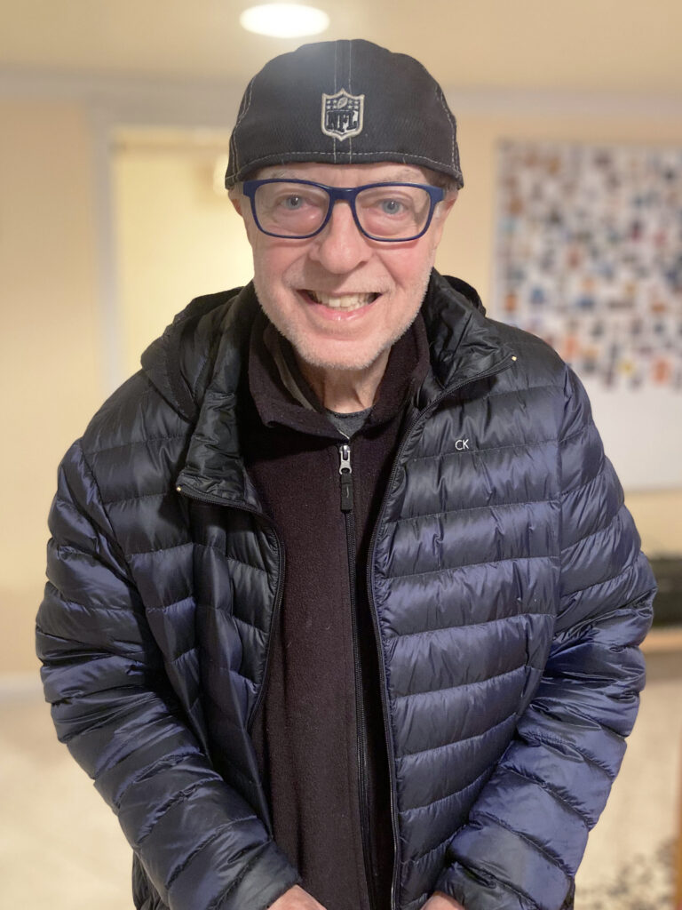 A man wearing glasses, a black cap, a black puffer jacket, and a dark shirt smiles while standing indoors.
