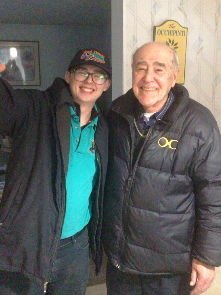 Two people stand indoors, both wearing winter jackets and smiling at the camera; one person is wearing a baseball cap and glasses.