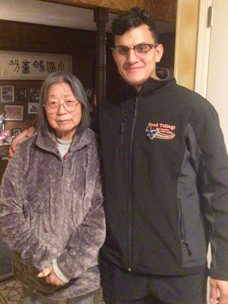 An older woman and a younger man stand side by side indoors, both facing the camera and smiling slightly.