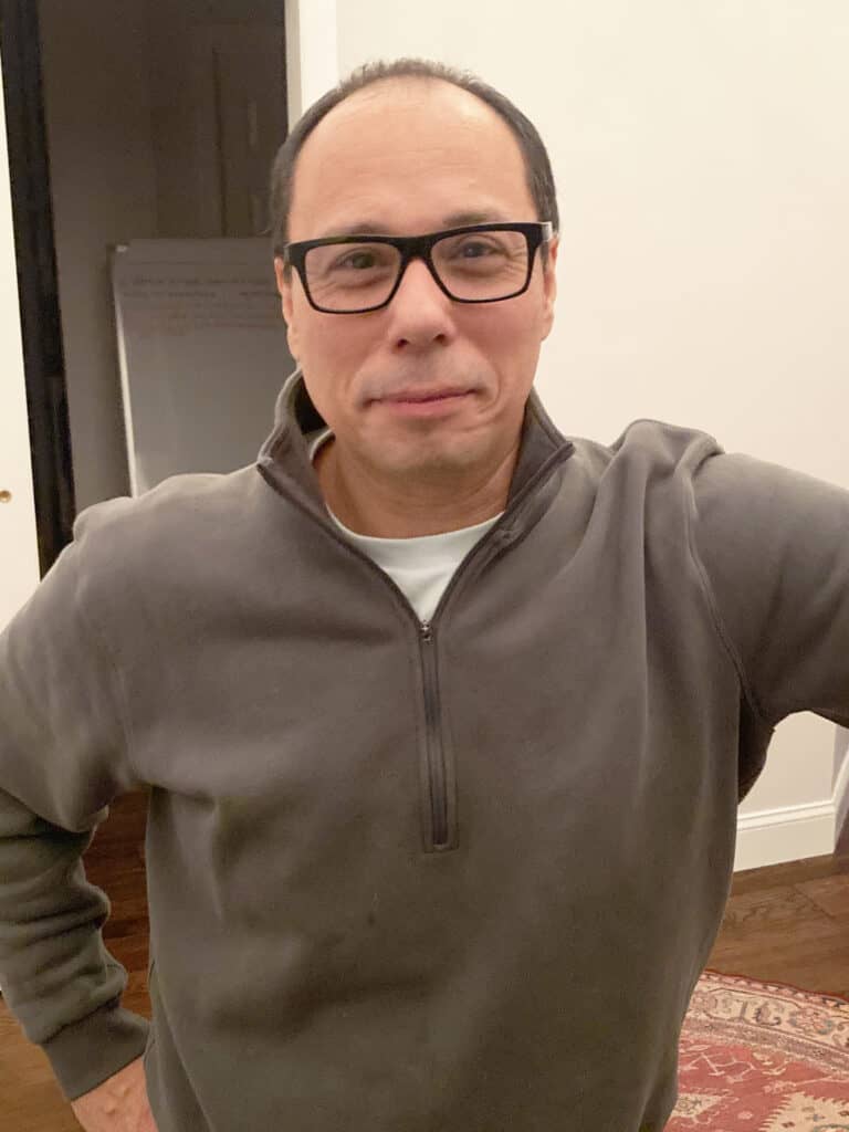 A man wearing glasses and a gray zip-up sweater stands indoors with one hand on his hip, facing the camera.