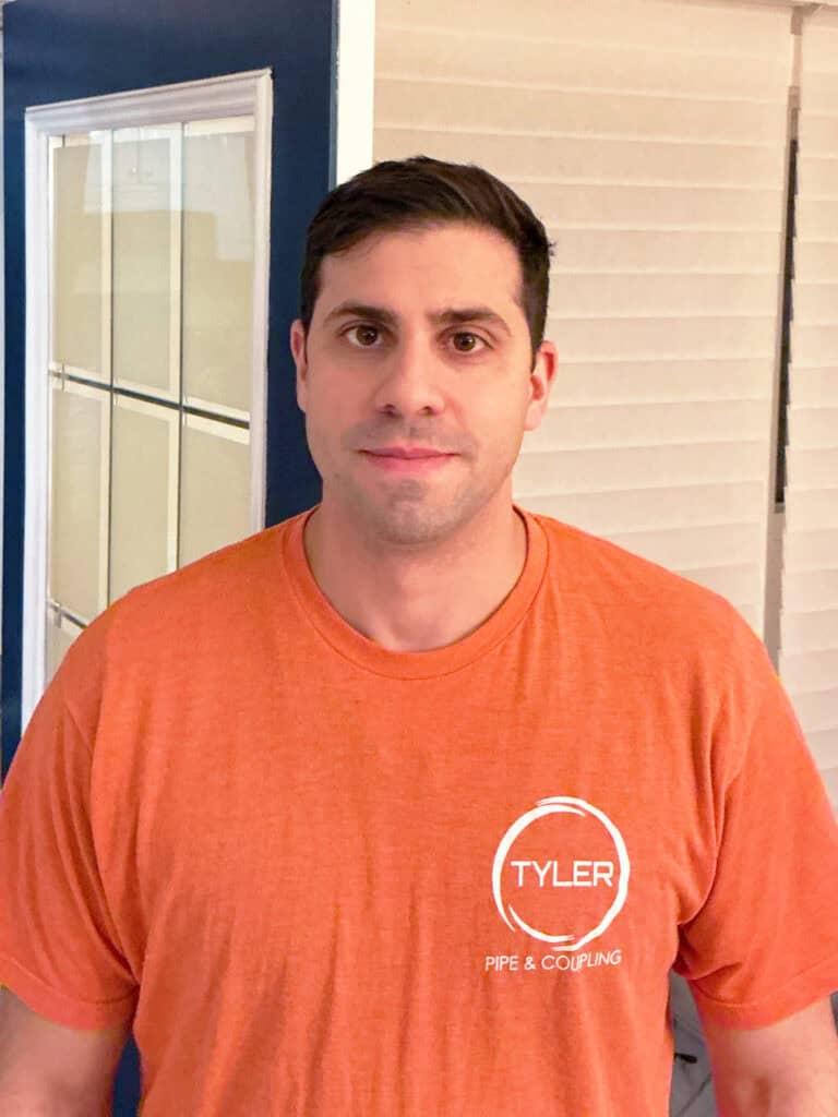 A man stands indoors, wearing an orange Tyler Pipe & Coupling T-shirt, in front of a blue door with glass panels and closed blinds.