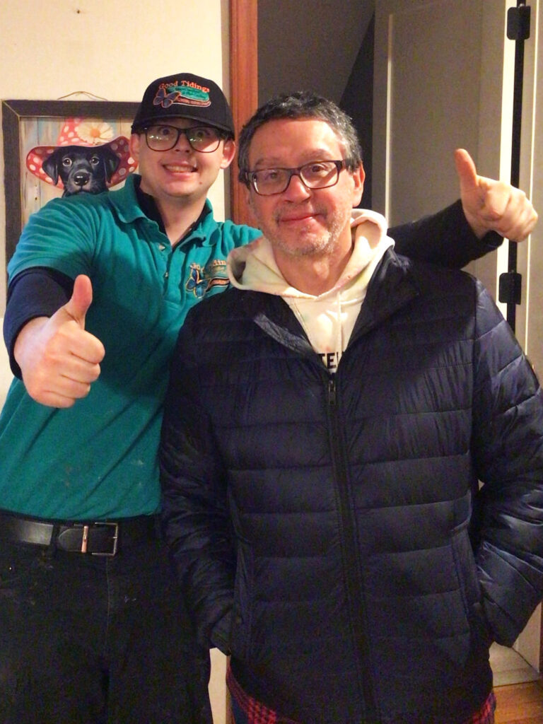Two men pose indoors; one in a green shirt and cap gives a thumbs up, the other wears glasses and a black jacket. A framed dog portrait hangs on the wall behind them.