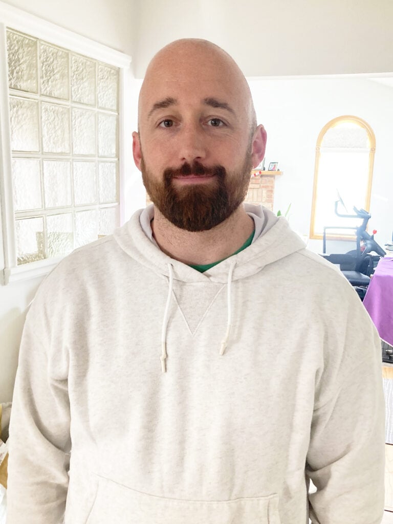 A man with a shaved head and beard is standing indoors, wearing a white hoodie and smiling slightly at the camera.