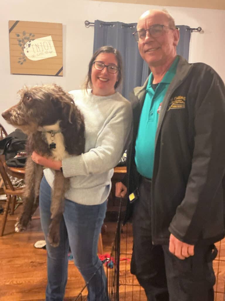 A woman holding a large dog stands next to an older man; they are indoors with furniture and a sign visible in the background.