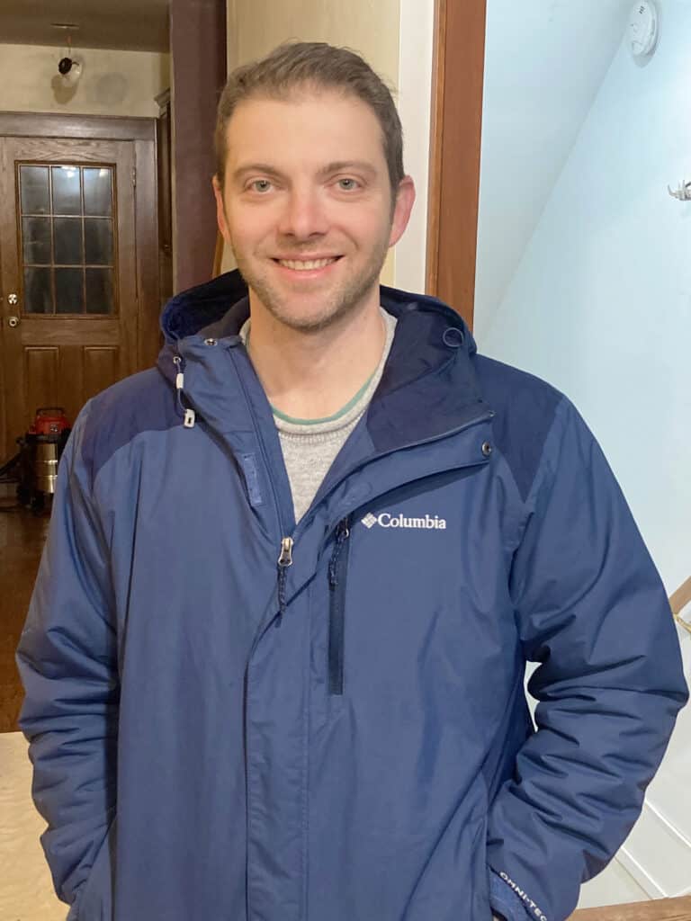 A man wearing a blue Columbia jacket stands indoors, smiling at the camera with one hand in his pocket. A vacuum is visible in the background near a wooden door.