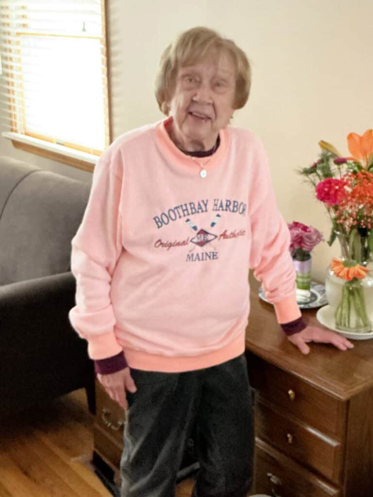 An elderly woman wearing a pink Boothbay Harbor Maine sweatshirt stands beside a wooden dresser with a vase of flowers in a living room.
