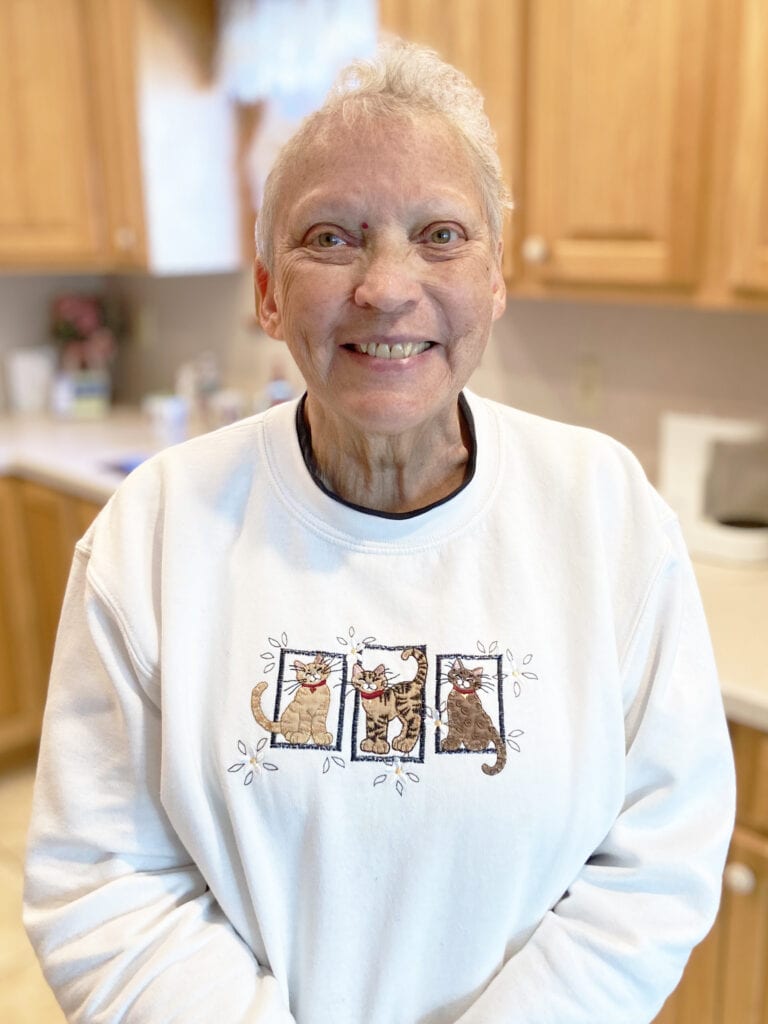 Smiling older woman with short gray hair stands in a kitchen, wearing a white sweatshirt with three cartoon cats on the front.