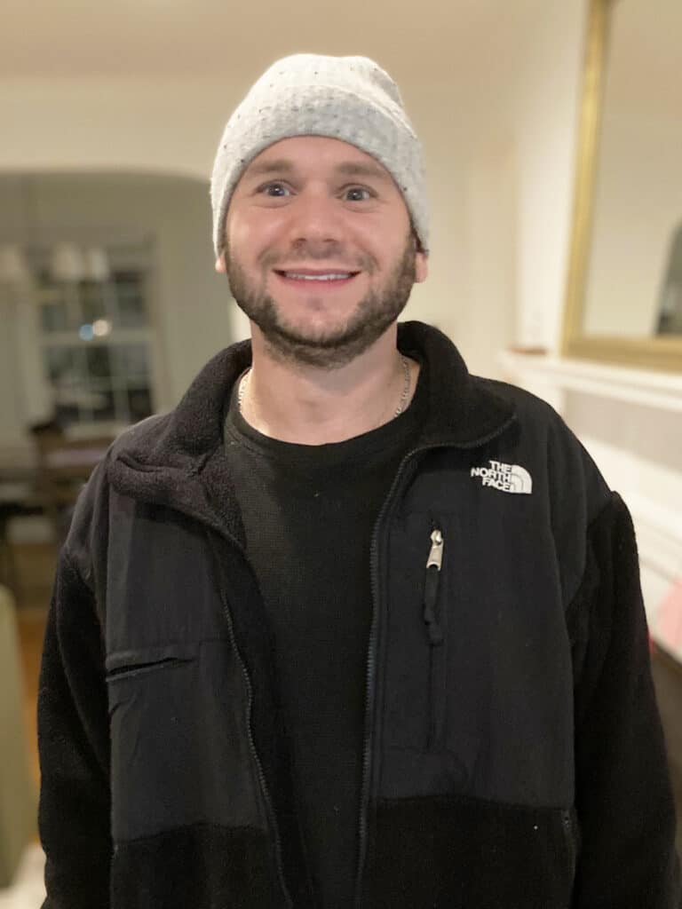 A man wearing a gray knit beanie, black North Face jacket, and black shirt stands indoors, smiling at the camera.