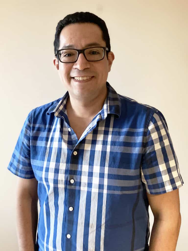 A man with short dark hair and glasses is smiling, wearing a blue and white plaid short-sleeve shirt, standing against a plain light-colored background.