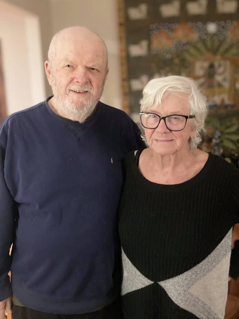 An older man and woman stand side by side indoors, both smiling at the camera. The background shows a patterned wall hanging.
