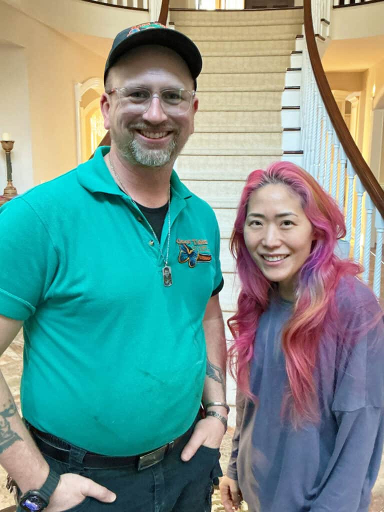 A man in a green shirt and a woman with long pink hair stand smiling indoors in front of a staircase.