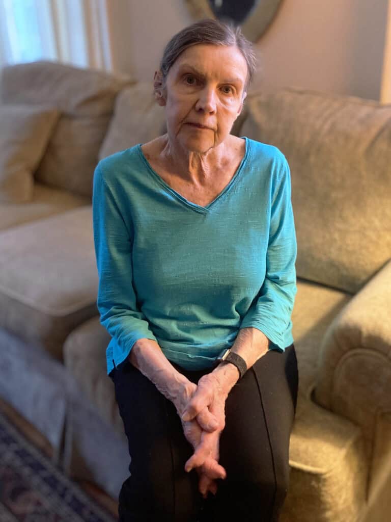 An older woman with gray hair sits on a beige couch, wearing a teal top and black pants, with her hands clasped in her lap.