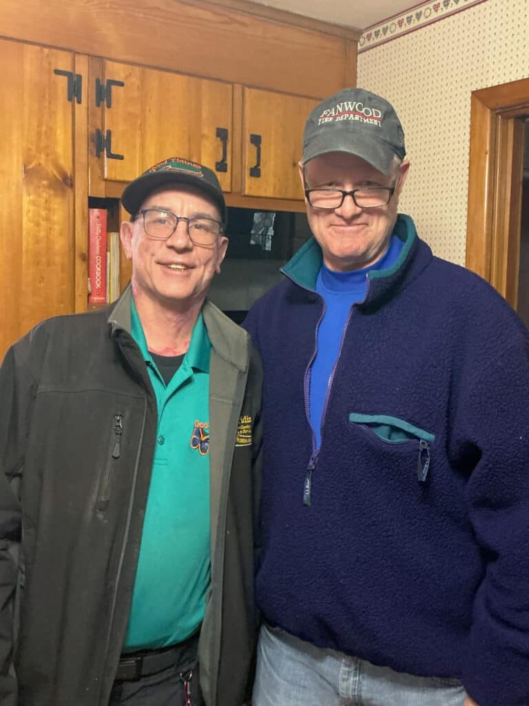 Two older men wearing glasses and hats stand side by side in a kitchen, smiling at the camera. One wears a green shirt and jacket; the other wears a blue fleece.