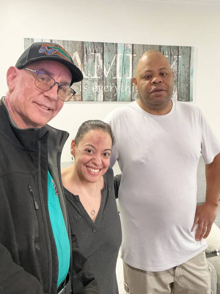 Three adults stand indoors, smiling at the camera. One man wears glasses and a cap, the woman is in the center, and another man in a white t-shirt is on the right.
