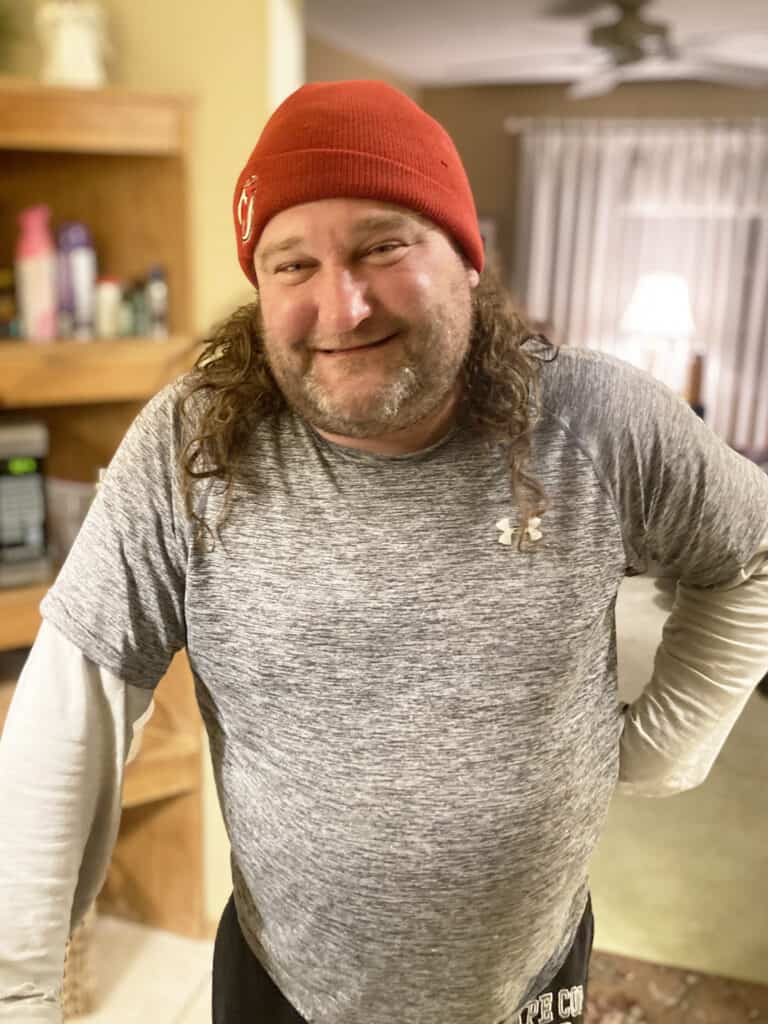 A man with long hair and a beard wearing a red beanie, a gray Under Armour shirt, and black pants stands indoors, smiling at the camera.